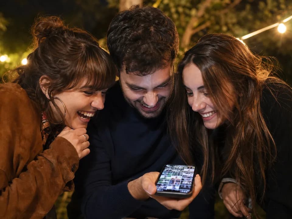 Drei Personen schauen lachend gemeinsam auf ein Smartphone mit einem Raster aus Fotos