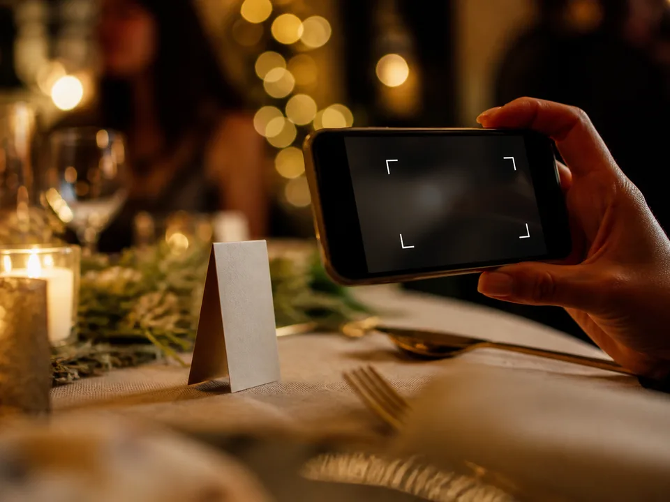 Eine Hand führt ein Smartphone an eine aufgestellte Tischkarte auf einem festlich gedeckten Tisch heran
